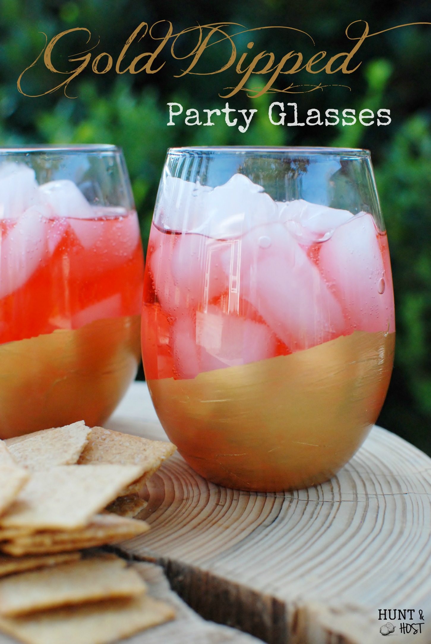 Two stemless glasses with painted gold bottoms filled with pink drinks and ice beside crackers on a wood slice