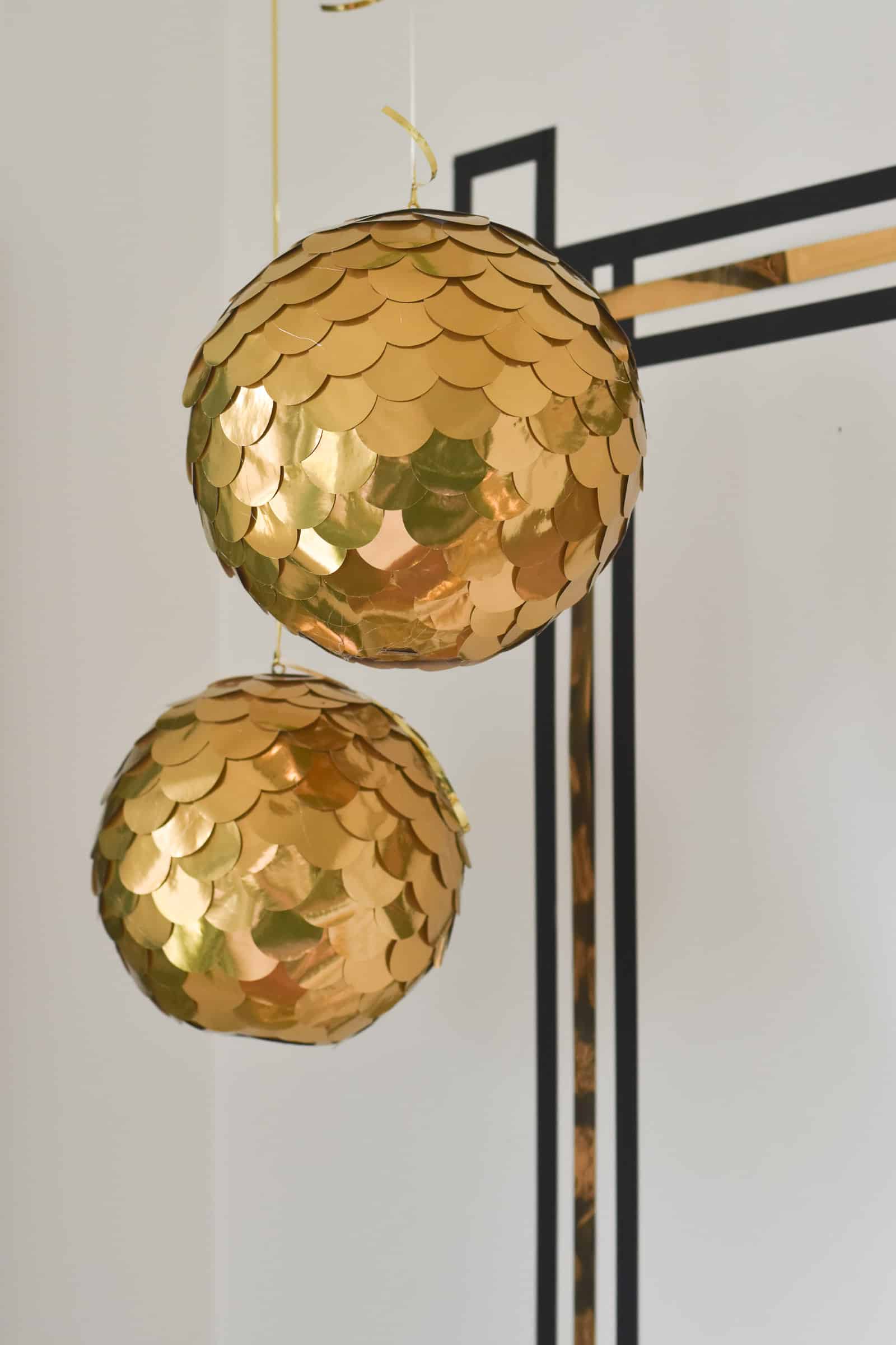 Two gold disco balls covered in overlapping metallic paper circles hanging on ribbon in front of a gold striped wall