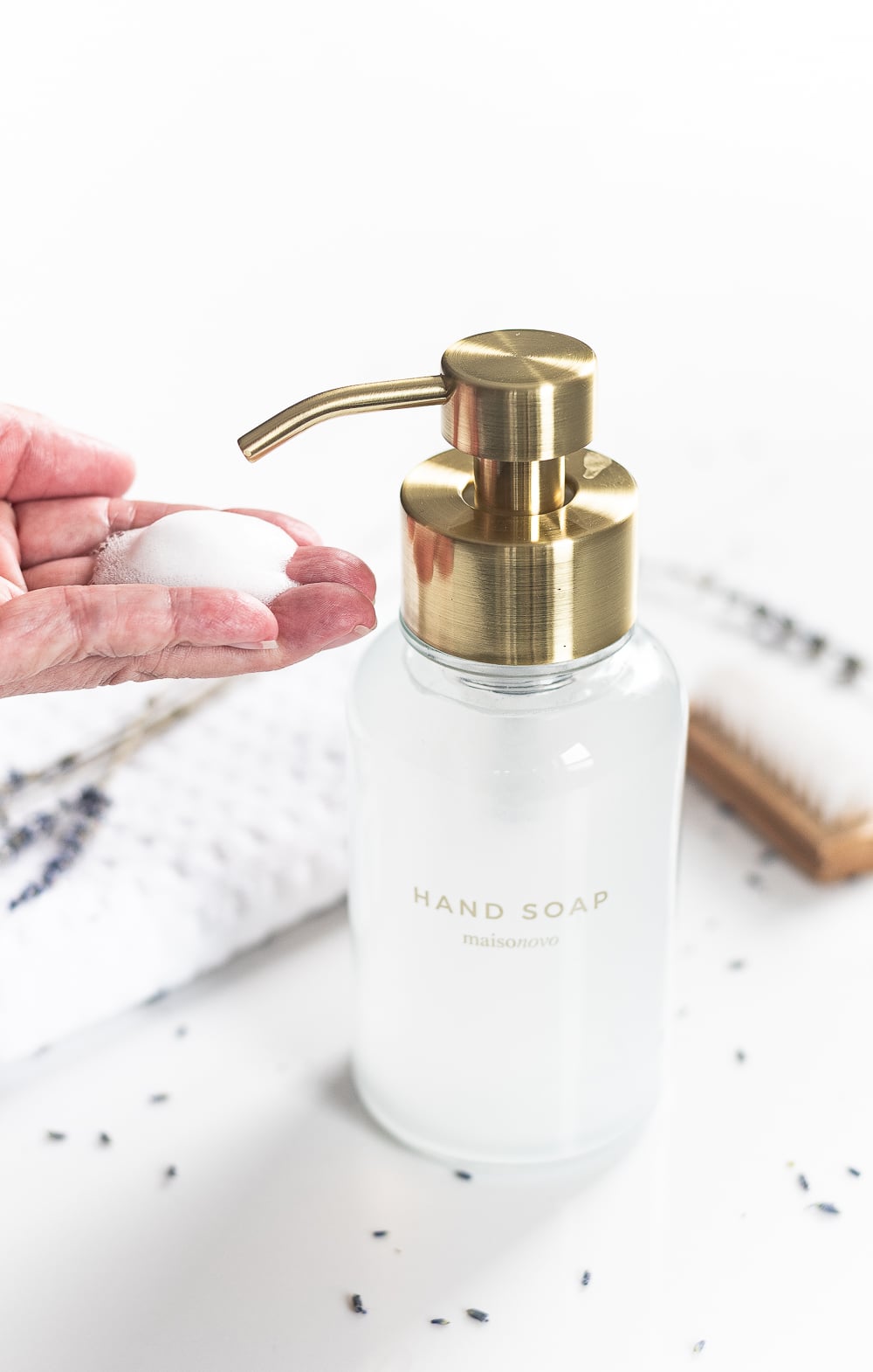 Frosted glass foaming soap dispenser with a brushed gold pump and a hand soap label as a hand catches the white foam