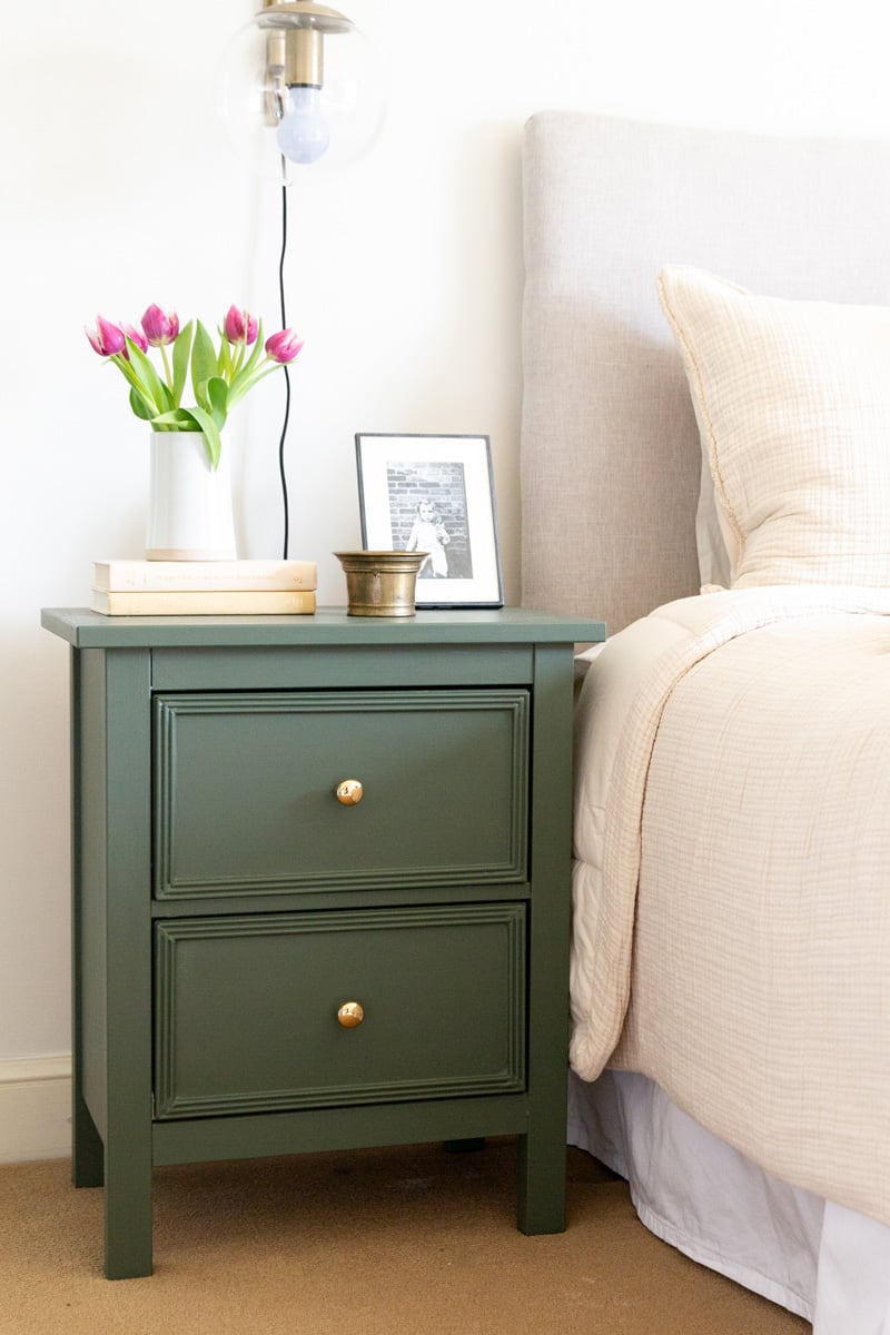 Green IKEA Hemnes Nightstand Makeover | IKEA Bedroom Hack IKEA Hemnes nightstand makeover painted deep green with framed drawer fronts and brass knobs, topped with pink tulips, books and a framed photo beside an upholstered bed