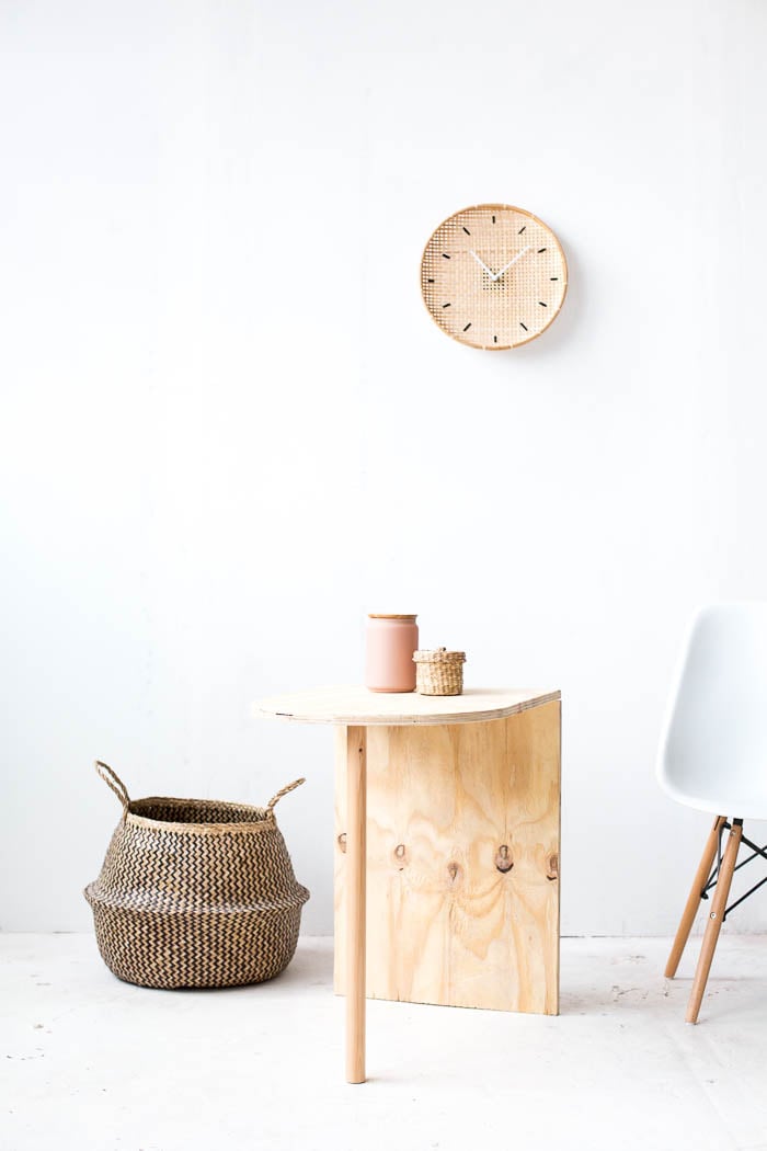 Light plywood side table with a rounded half circle top and one dowel leg beside a woven basket and a white chair