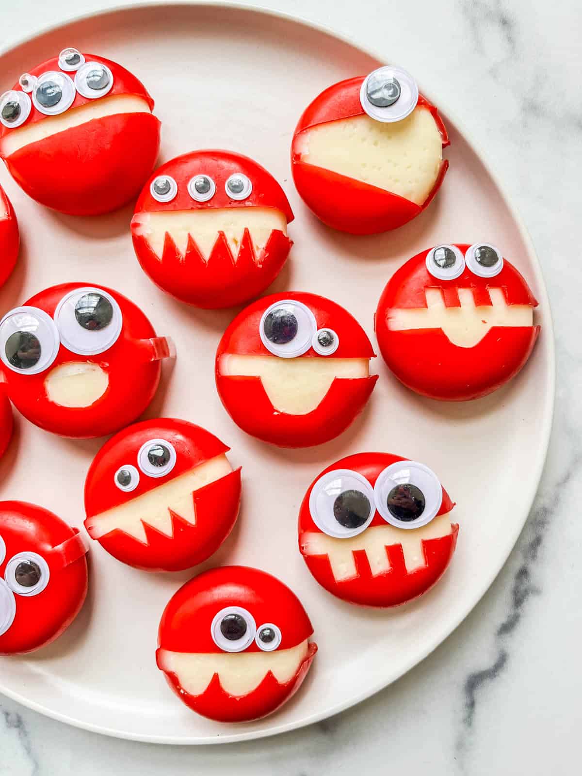 Red wax Babybel cheese rounds with zigzag mouths cut to show the cheese and googly eyes, on a white plate on marble