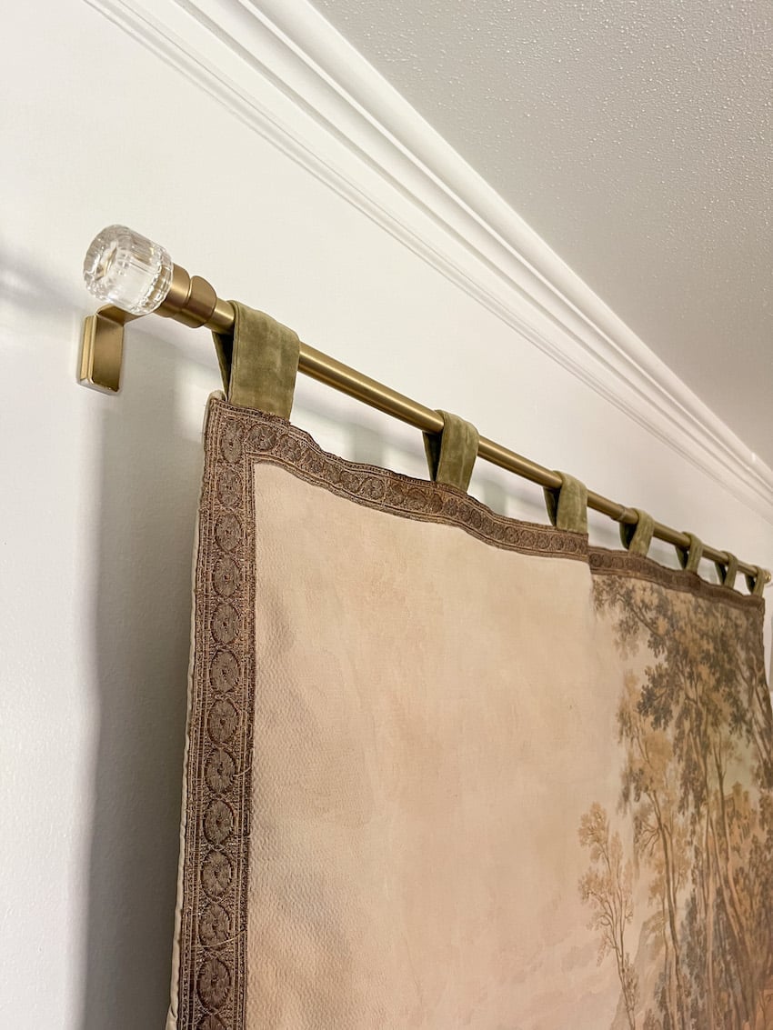 Vintage woven landscape tapestry with tab tops hanging from a brass curtain rod with a clear glass finial on a wall