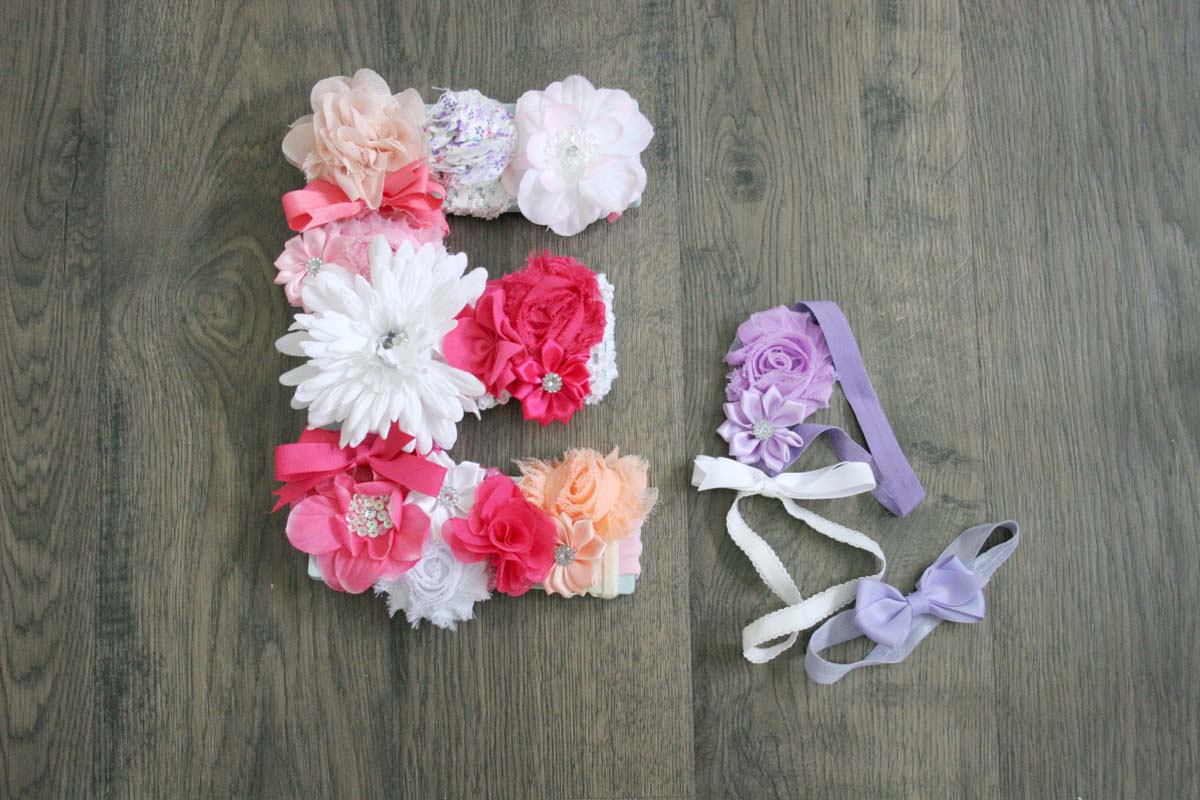 Wooden letter covered in pink, peach and white baby headband flowers on a gray wood floor beside lavender floral headbands