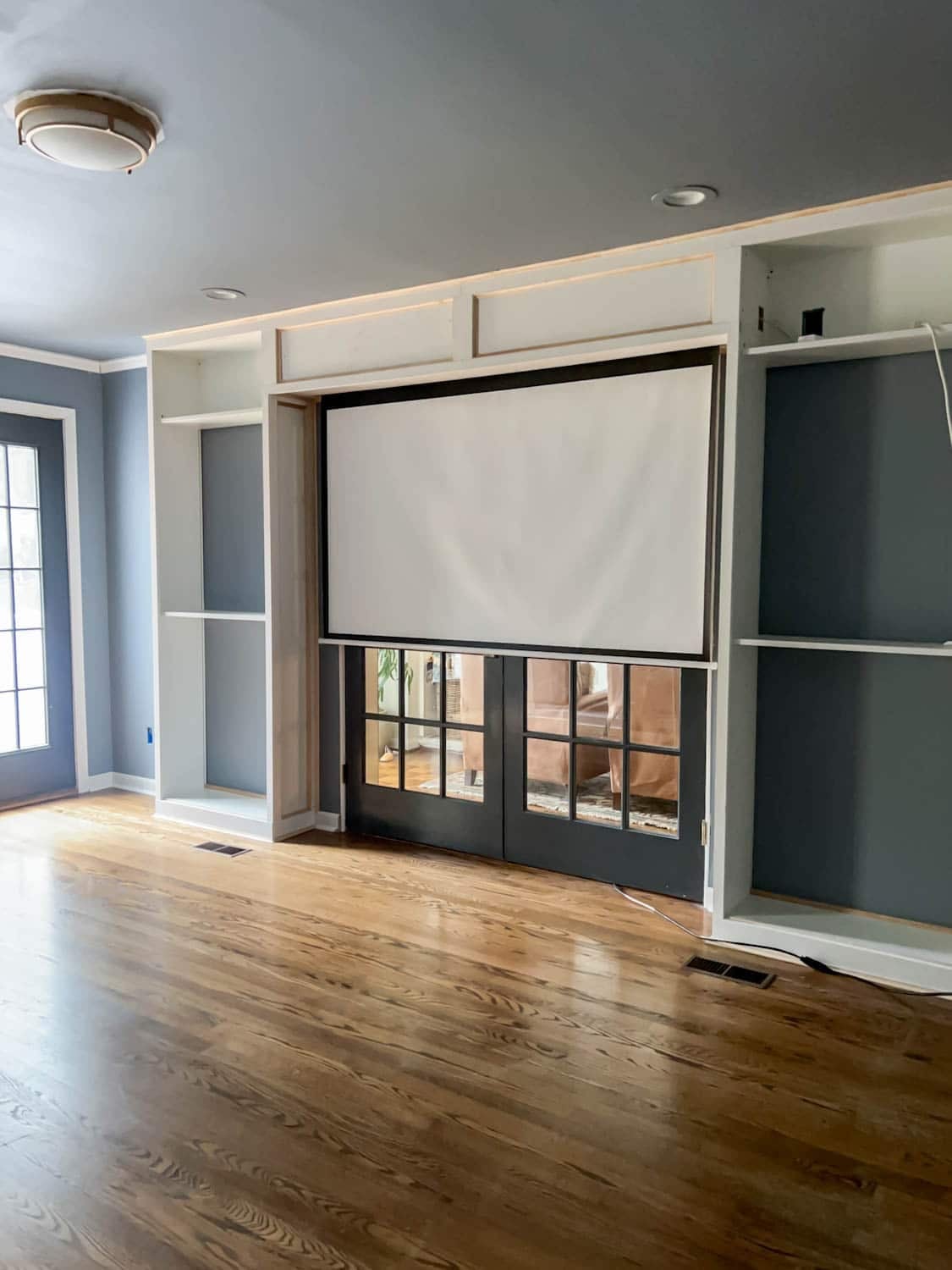 Hidden Projector Screen IKEA Hack | DIY Media Room Idea White built-in bookcases framing French doors with a pulled down projector screen hidden in a trimmed box above them