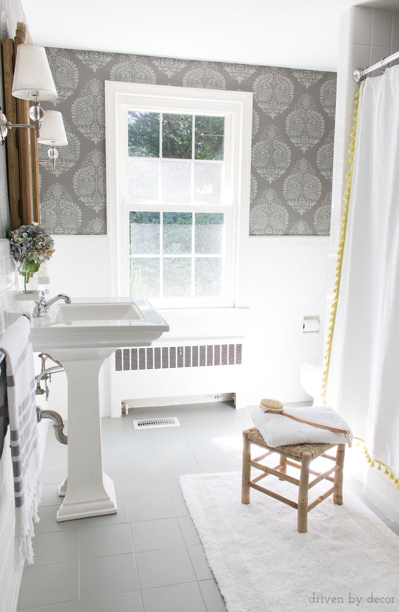 How to Paint a Tile Floor | Bathroom Floor Update Painted tile floor in soft gray in a white bathroom with a pedestal sink, wood stool, stenciled wallpaper and a white rug
