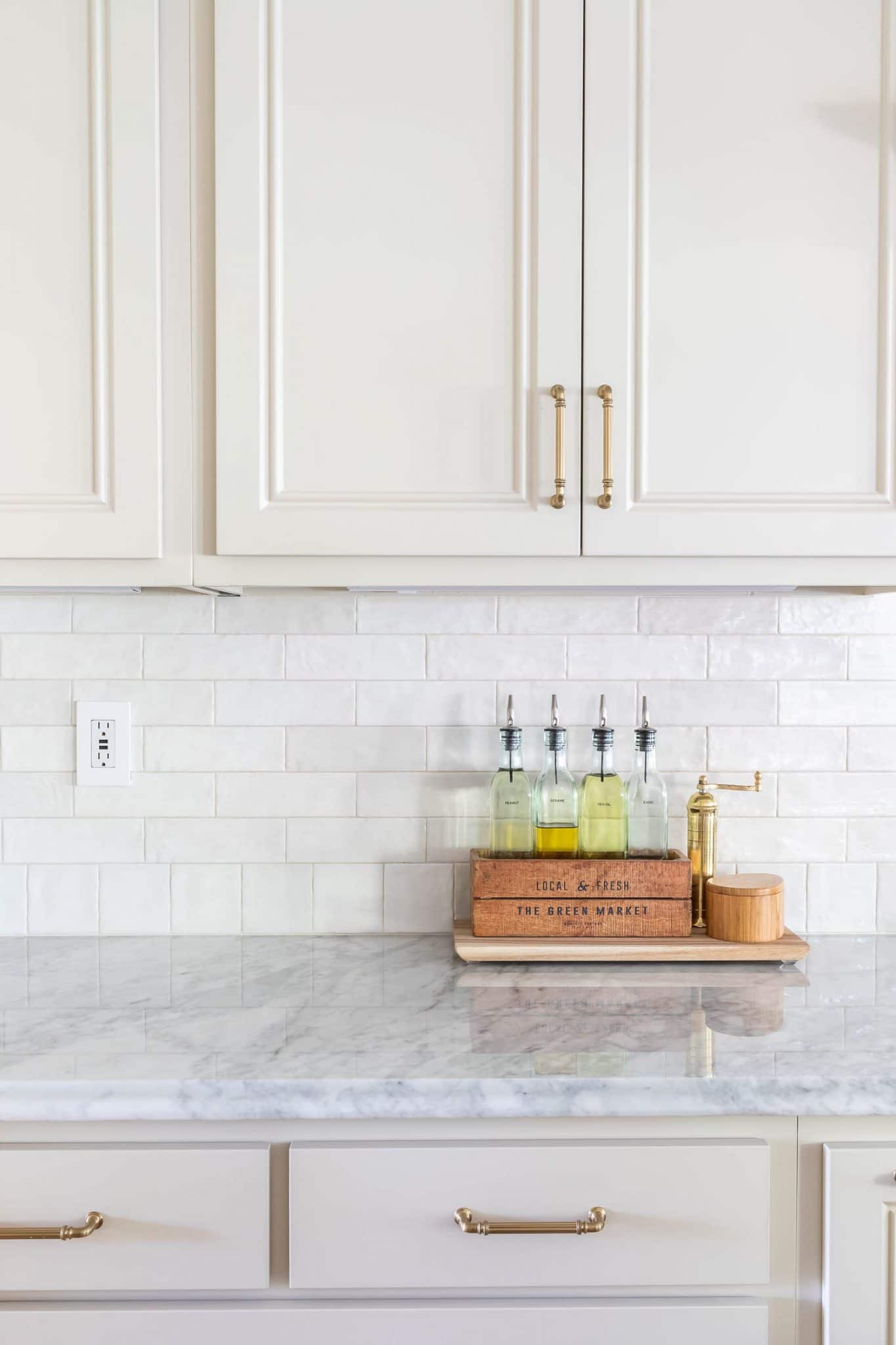 How to Tile a Backsplash | Easy Subway Tile Install White subway tile backsplash between cream kitchen cabinets with brass pulls, a marble counter and oil bottles on a tray