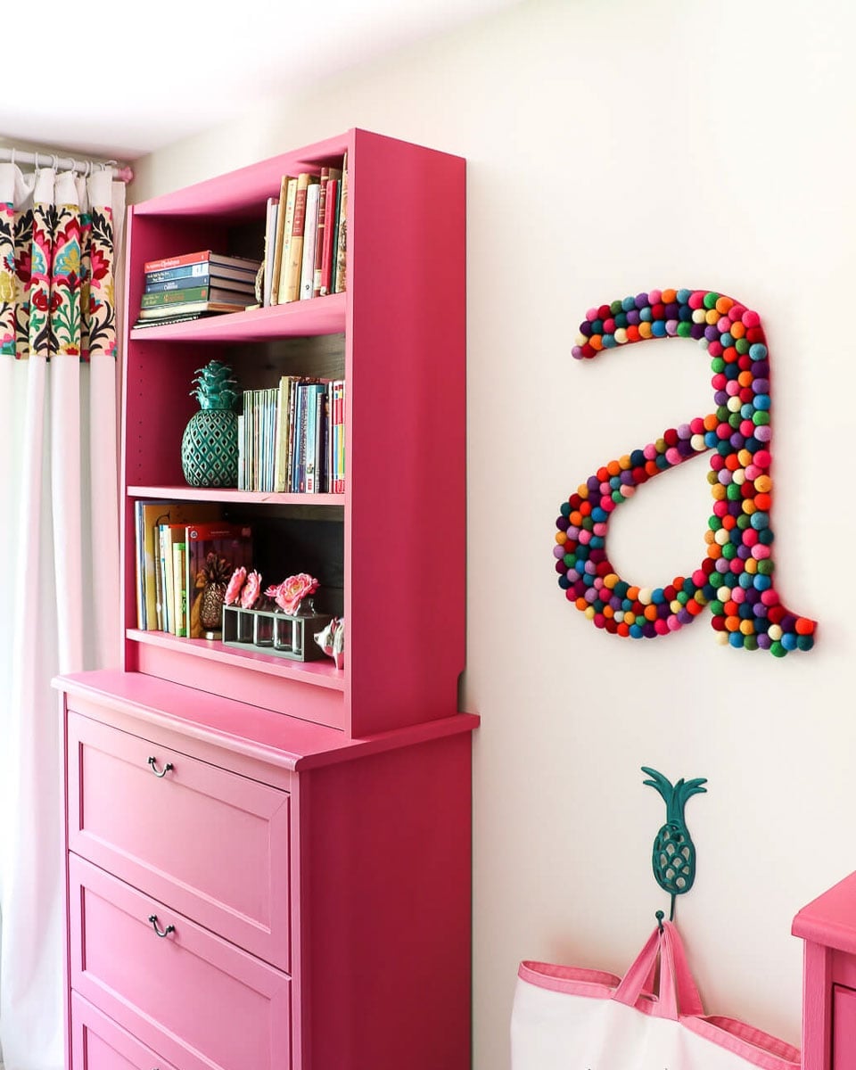 Hot pink painted IKEA Billy bookcase stacked on a matching dresser as a hutch, filled with books beside a felt ball letter