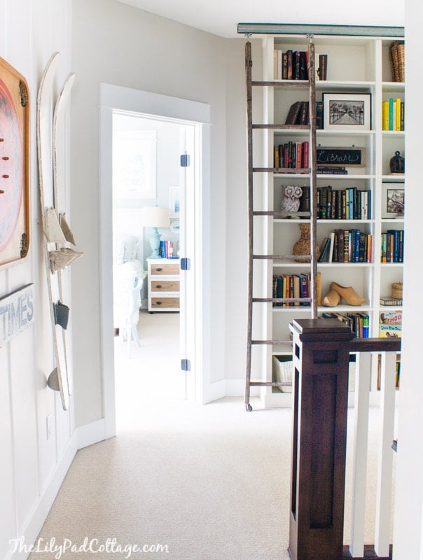 Floor to ceiling white IKEA Billy bookcases full of books with a rustic wooden library ladder hanging from a metal rail