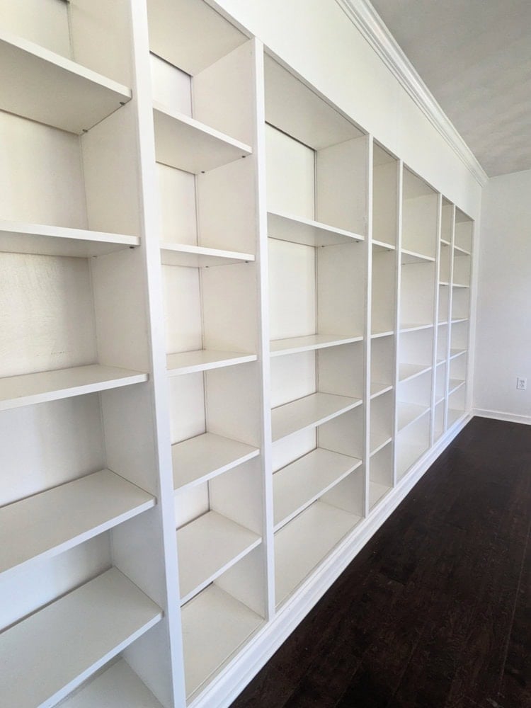 Long wall of white IKEA Billy bookcases joined with trim and crown molding into built-in shelves above a dark wood floor