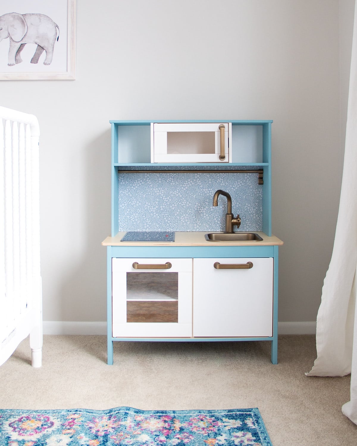 IKEA Duktig play kitchen painted white and soft blue with patterned backsplash, brass faucet and handles beside a crib