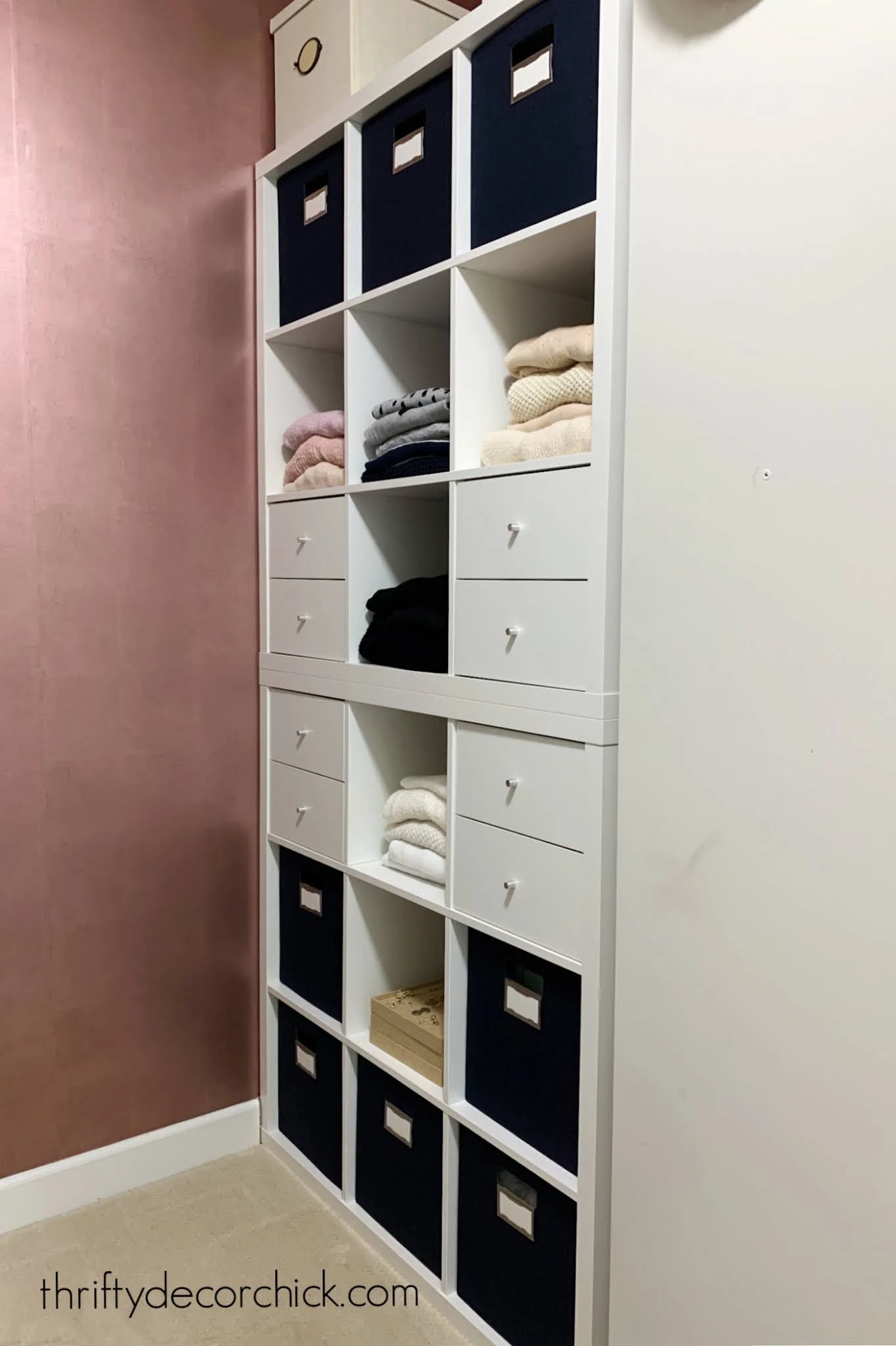 IKEA Kallax Closet Cubby Storage | IKEA Bedroom Hack Two stacked white IKEA Kallax cube shelves in a closet with navy bins, white drawers and folded sweaters by a pink wall