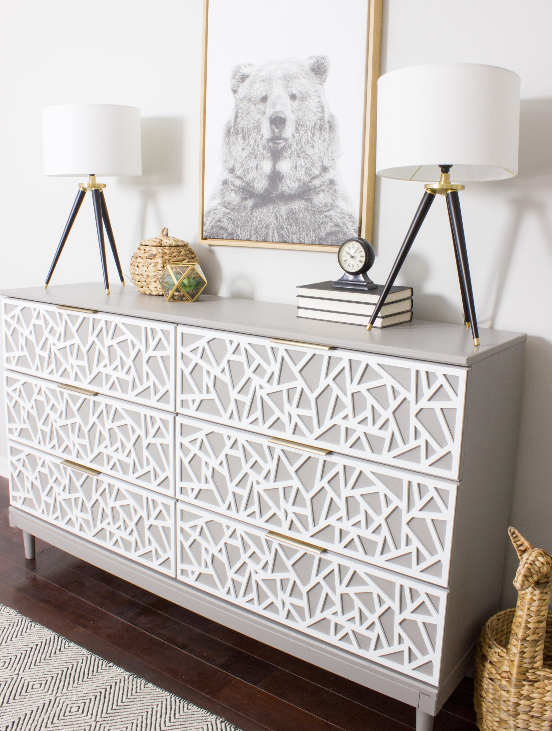 Gray IKEA TARVA dresser with white geometric overlays on six drawers, brass pulls and two tripod lamps under a bear print