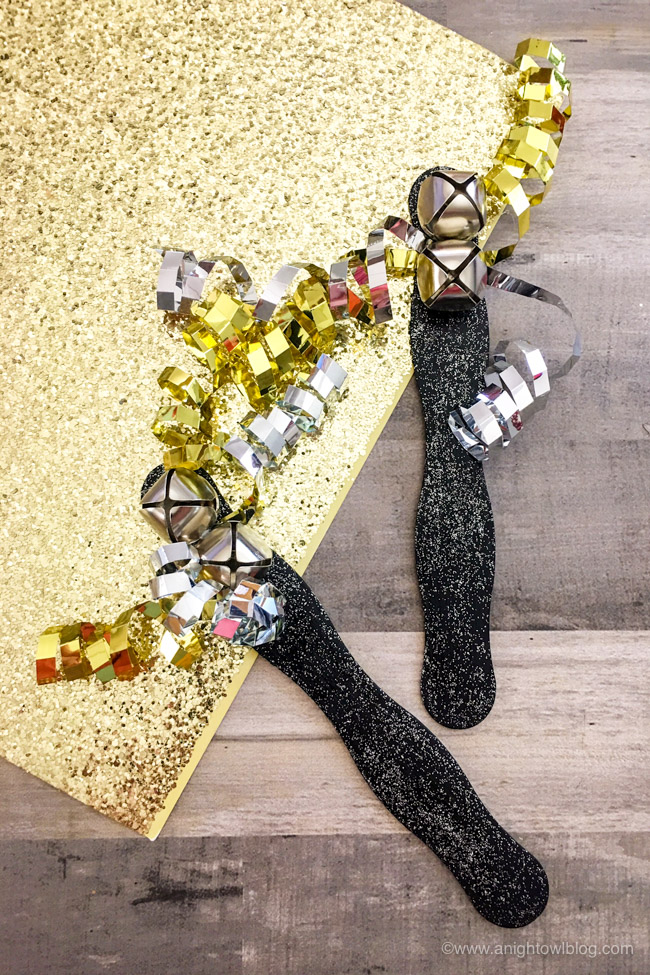 Two black glitter craft stick noisemakers with silver jingle bells and gold and silver curly ribbon on gold glitter paper