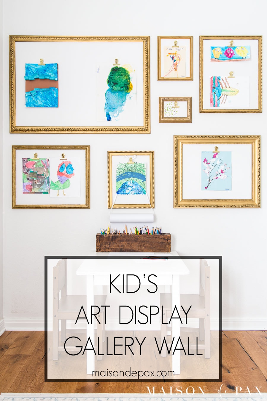Gallery wall of gold frames with clips holding colorful children's paintings above a small white kids table