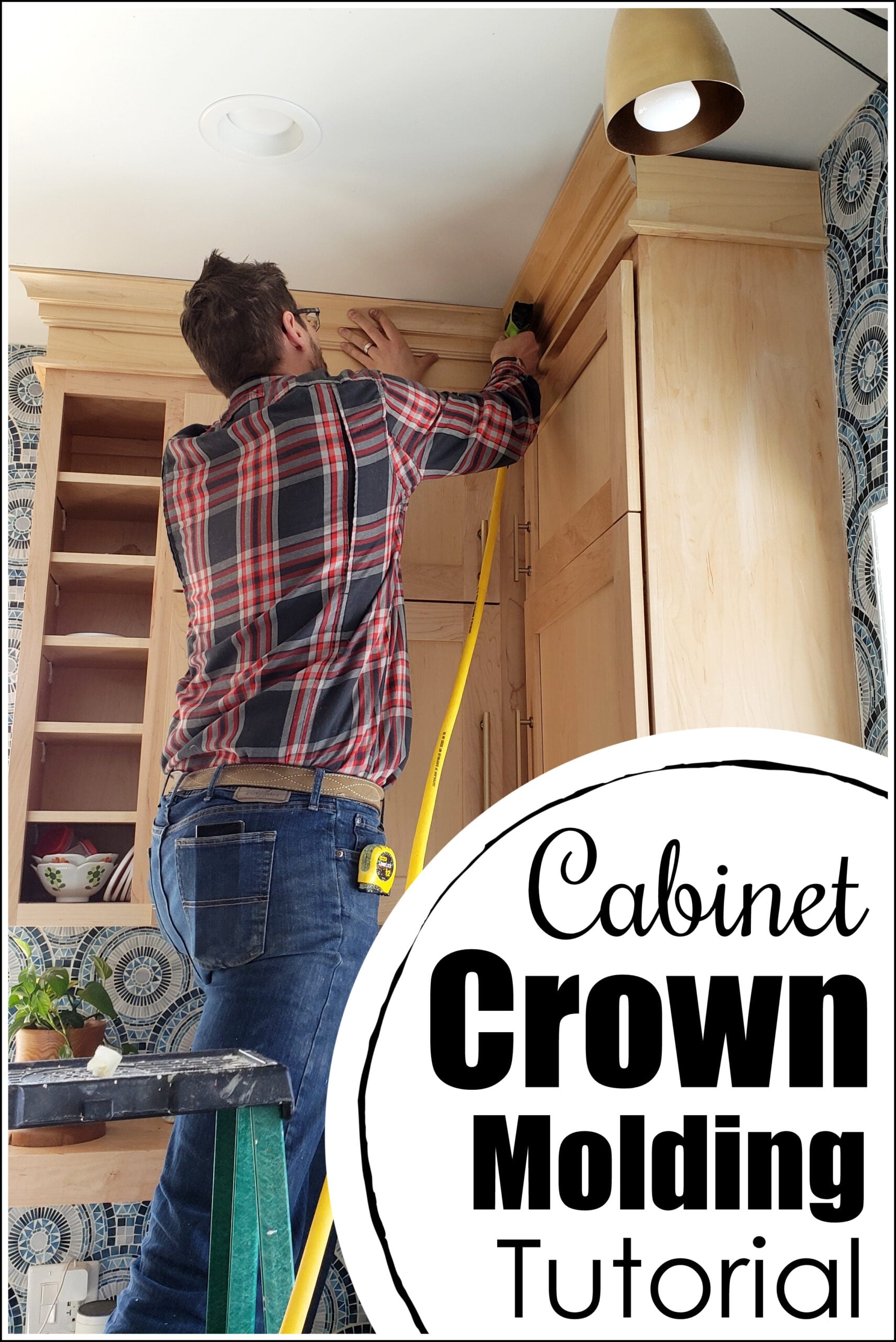 Man on a step stool fitting layered crown molding onto tall natural wood kitchen cabinets that reach a white ceiling