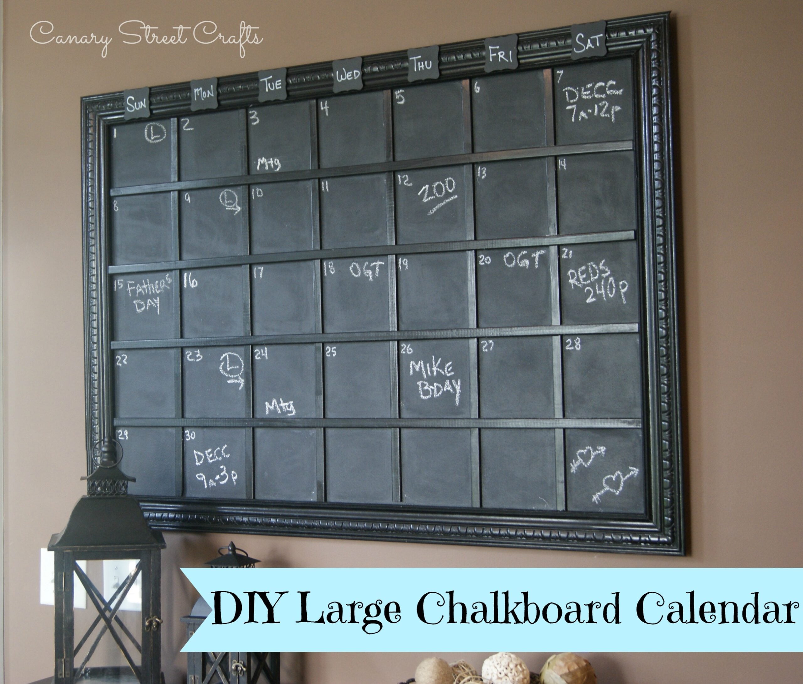 Large black framed chalkboard calendar with a moulding grid and day-of-week tags, hung above a lantern on a taupe wall