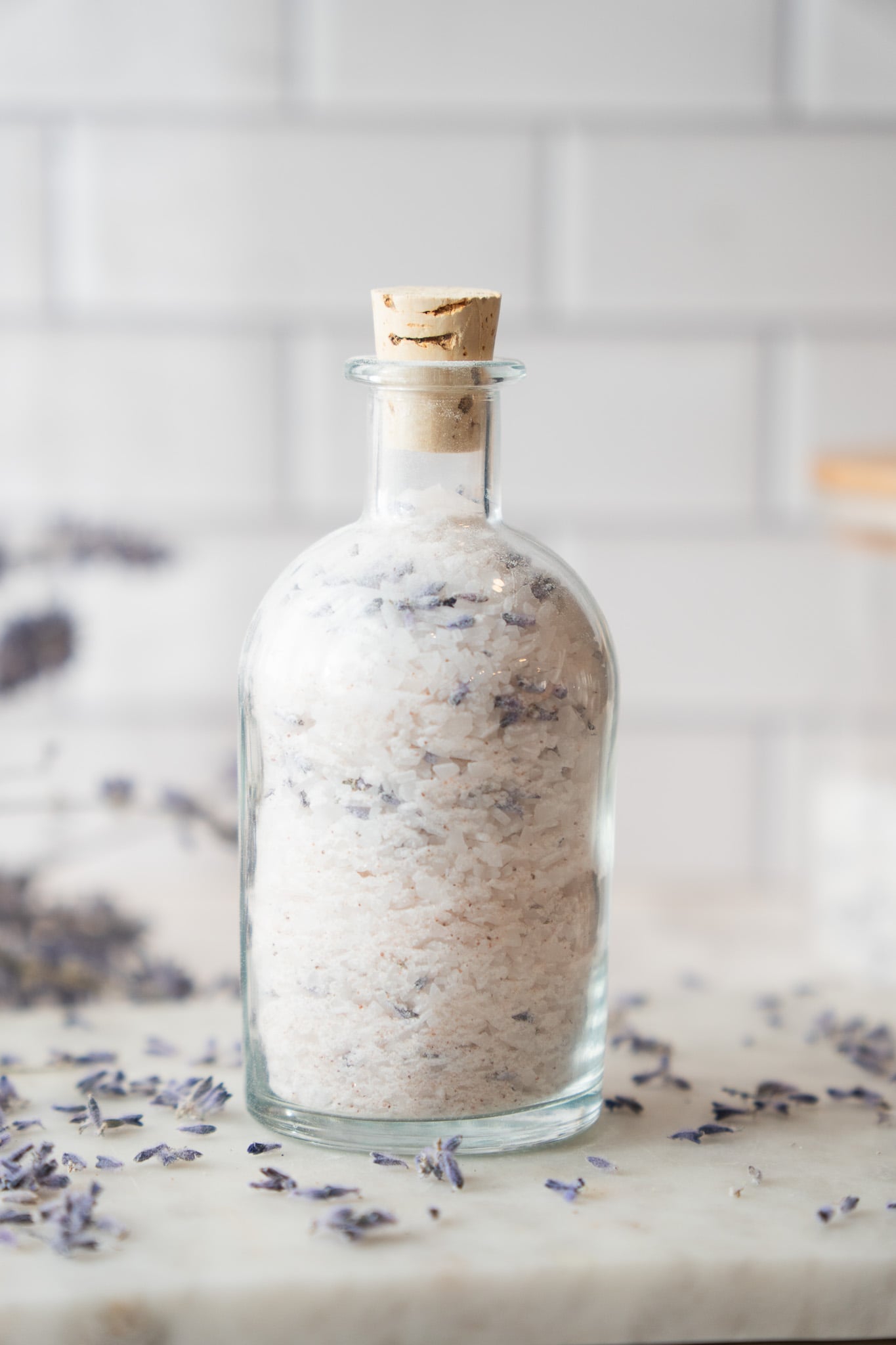 Corked glass bottle filled with homemade lavender bath salts on white marble with loose dried lavender buds around it