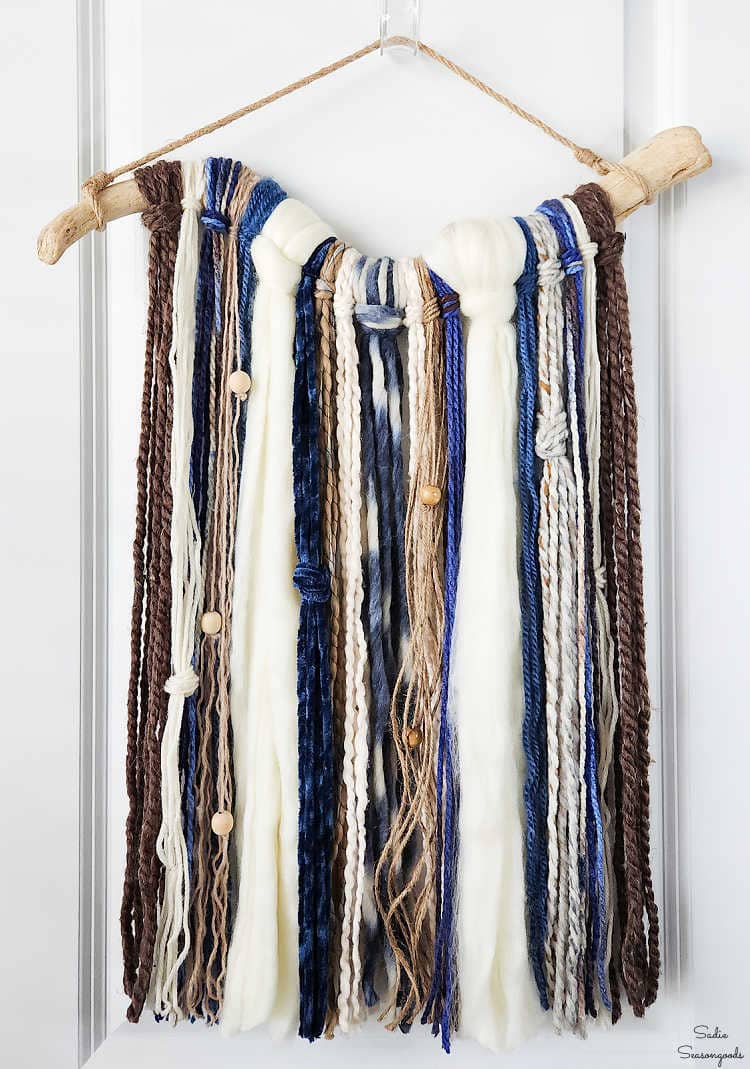 Boho yarn wall hanging of blue, cream, brown and gray strands with a few wooden beads hanging from driftwood on jute