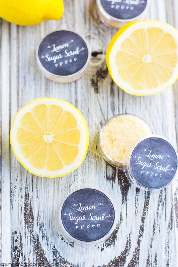 Small round tins labeled Lemon Sugar Scrub filled with homemade lip scrub beside fresh lemon halves on whitewashed wood