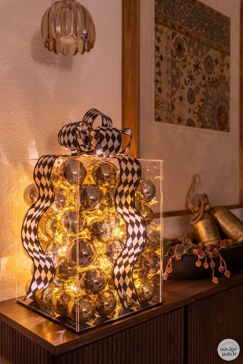 Light-Up Present Decor | DIY Party Decorations Idea Clear acrylic box filled with gold ornaments and glowing fairy lights, topped with a black and white harlequin ribbon bow