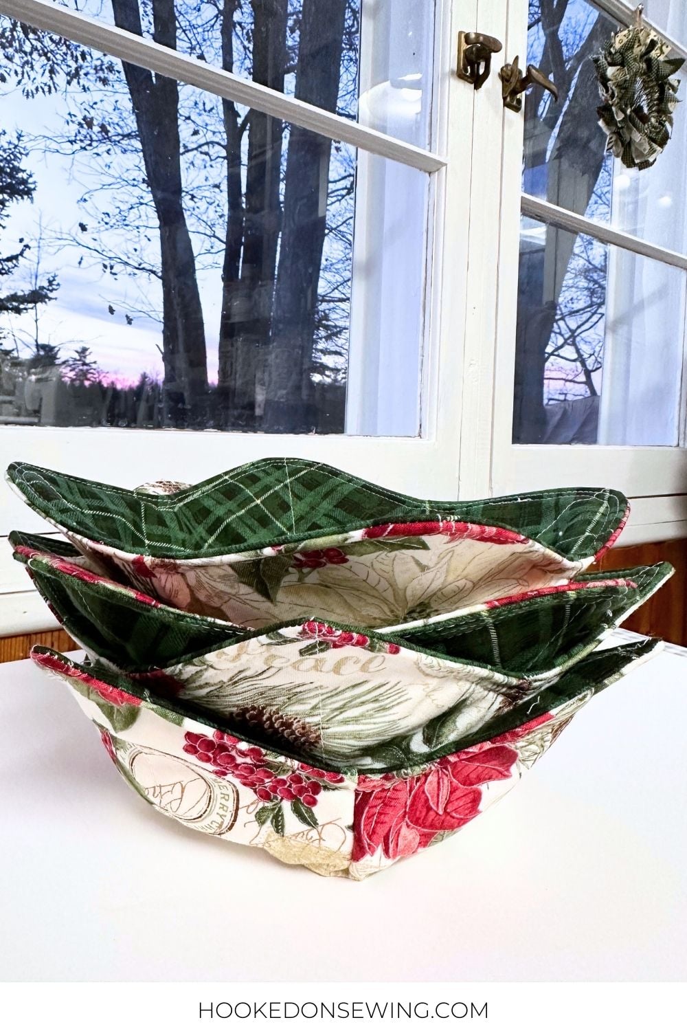 Microwave Safe Bowl Cozy Tutorial Stack of quilted bowl cozies in green plaid and red rose Christmas prints on a white counter by a snowy window