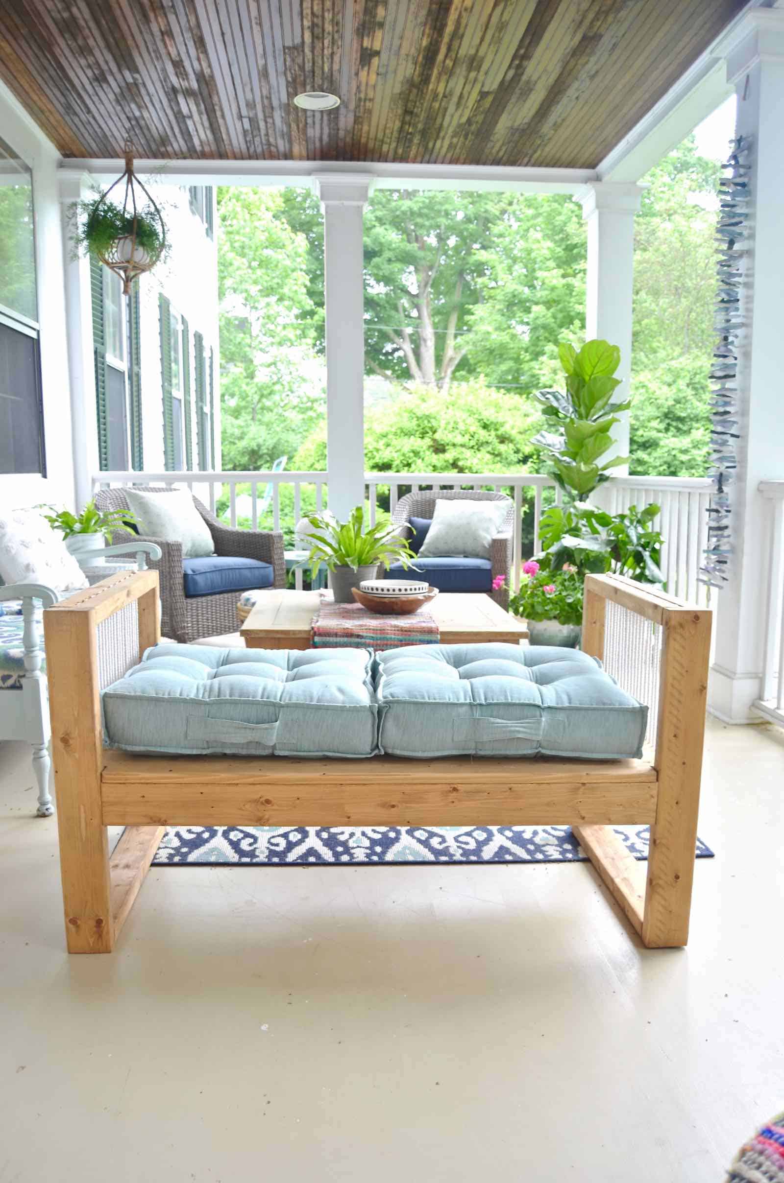 Mid Century Rope Bench | DIY Patio Furniture Idea Golden stained wood bench with white cotton rope through the arms and an aqua cushion on a covered porch with a rug