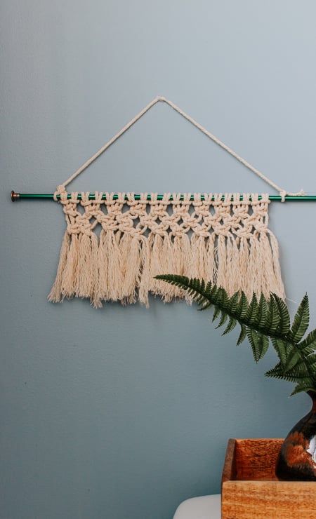 Small cream macrame wall hanging with rows of square knots and brushed fringe on a green knitting needle on a blue wall