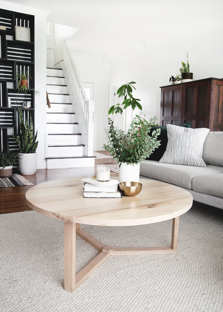 Large round light maple coffee table on a modern three leg wood base with plants, books and a gold bowl in a bright room
