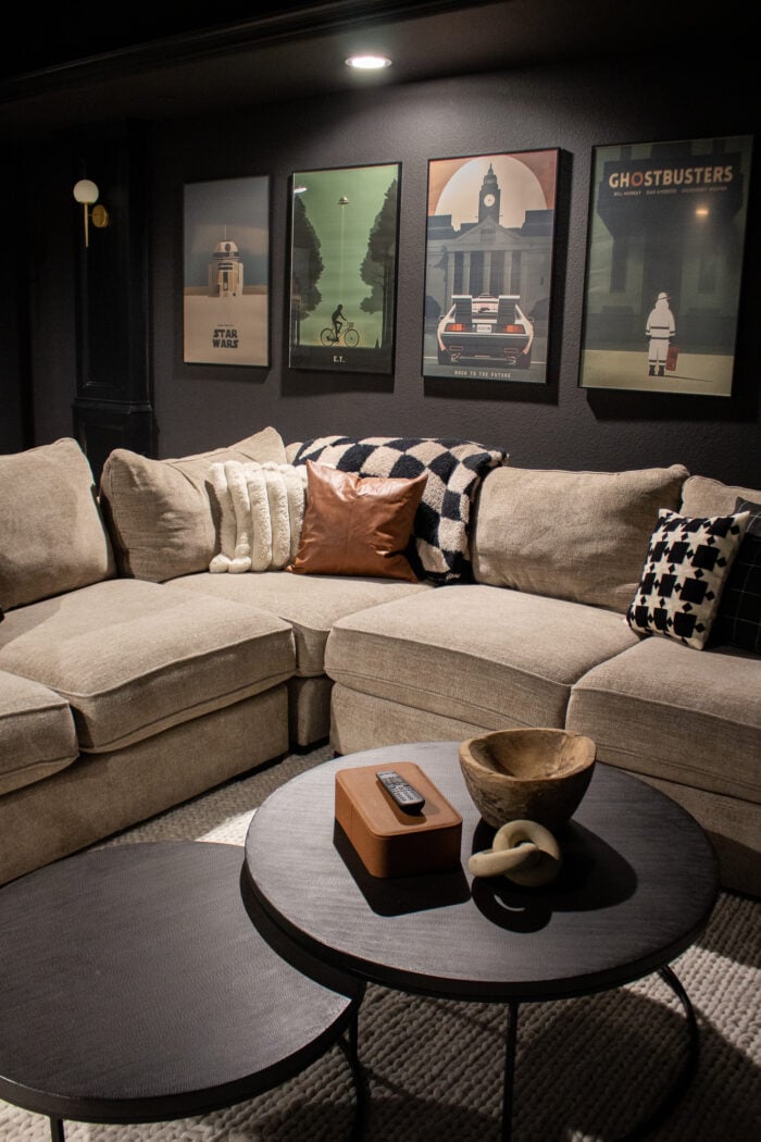 Moody Movie Poster Home Theater | DIY Media Room Idea Black home theater with a row of framed vintage style movie posters above a sectional sofa piled with cream and black pillows