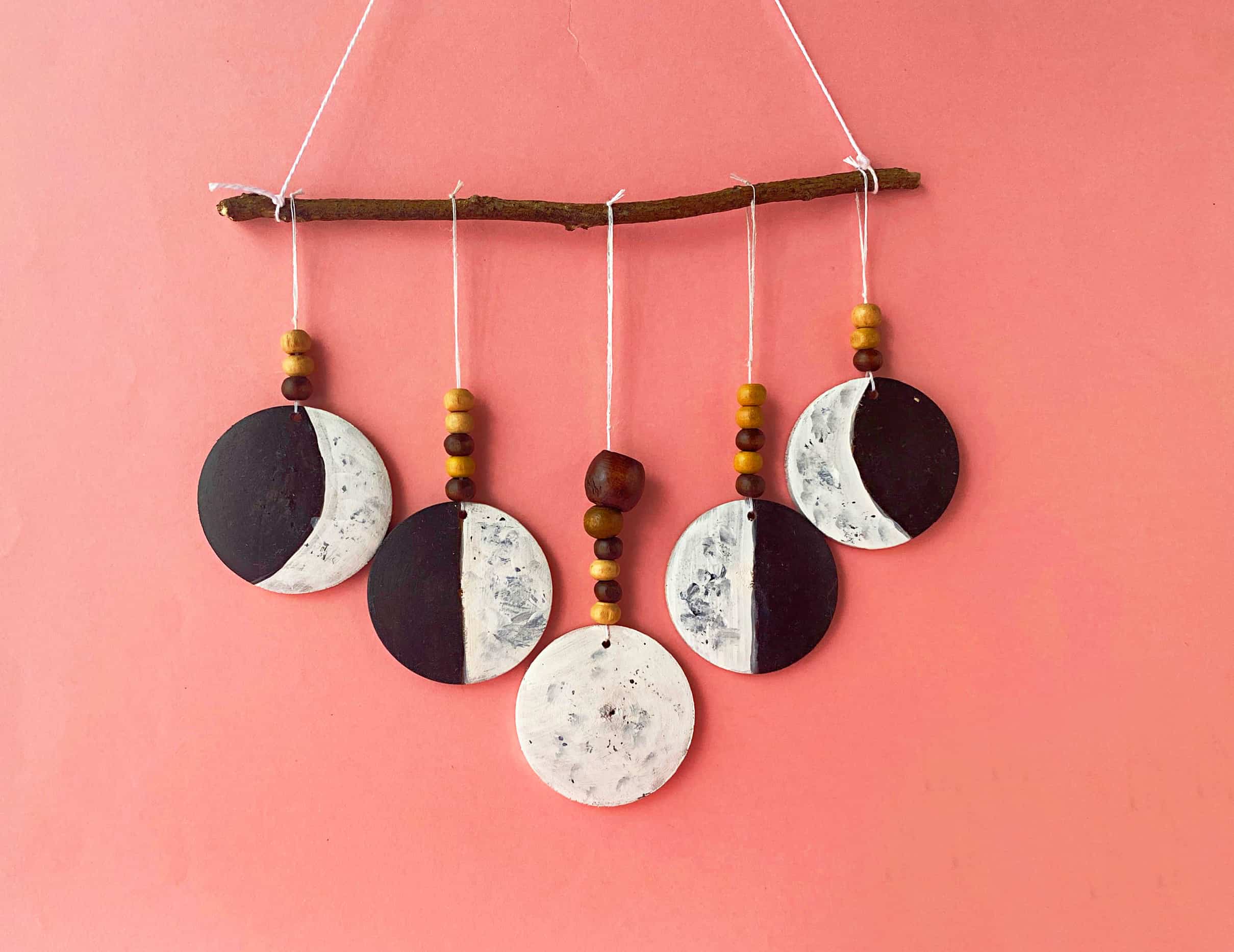 Five painted wood discs showing moon phases in black and white strung with wooden beads from a twig on a coral pink wall