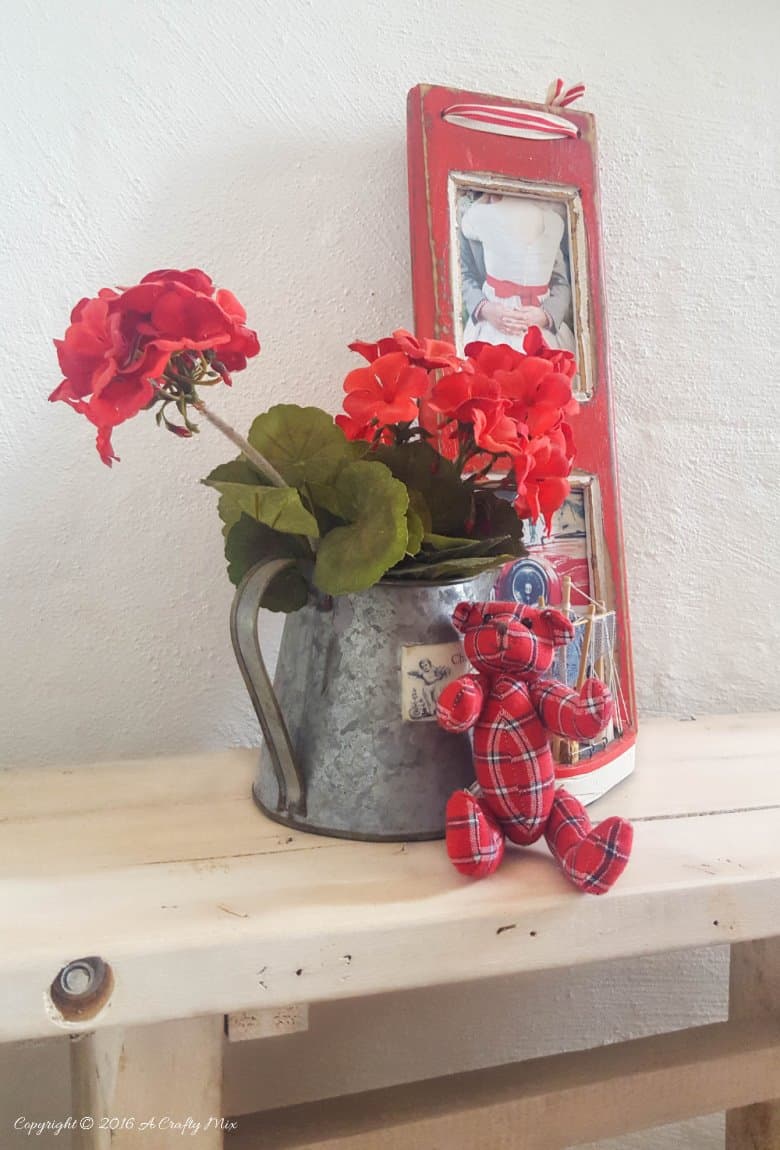 Narrow whitewashed pallet wood side table in a white corner holding a watering can of red geraniums and a red photo frame