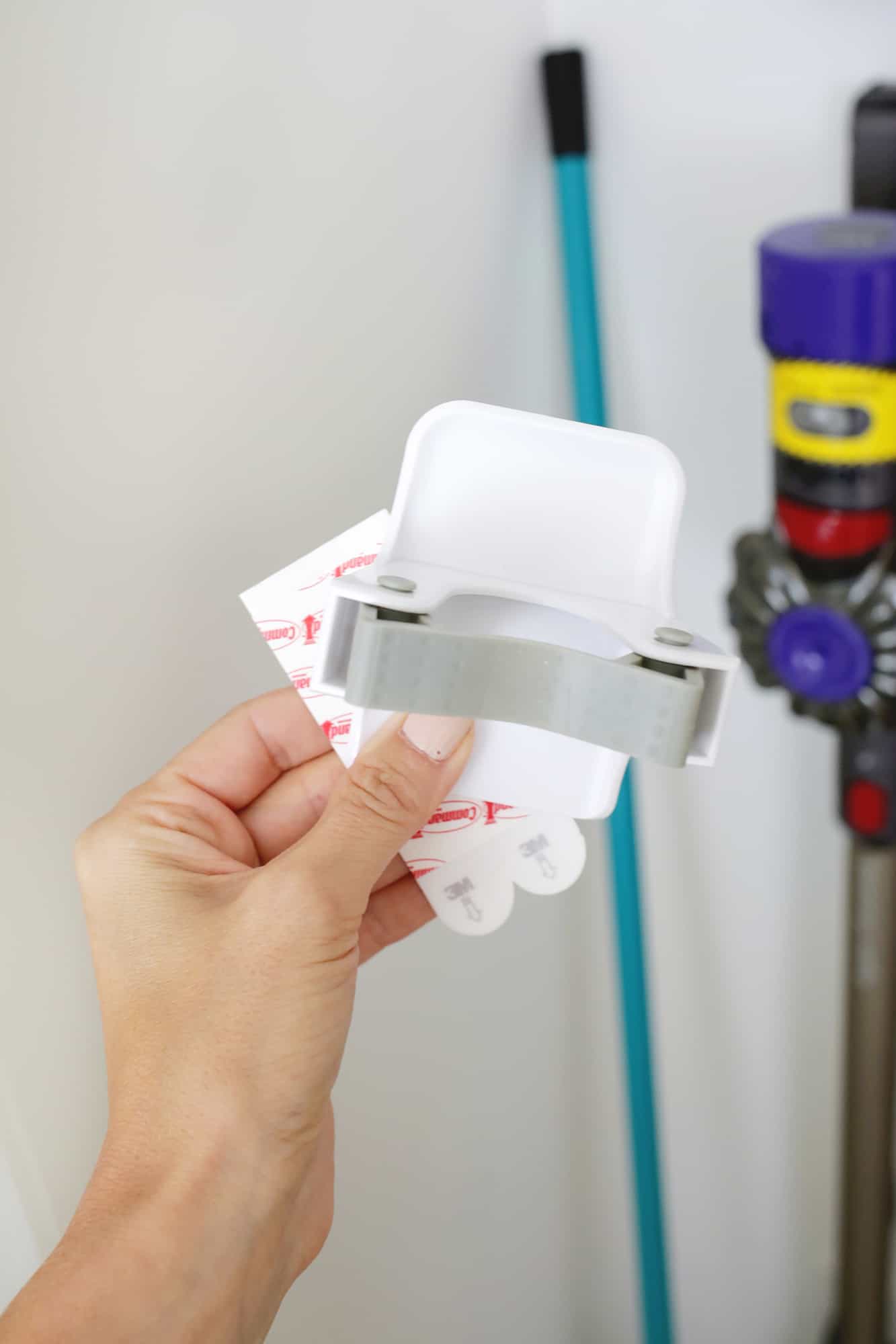 Hand holding a white adhesive broom grip hook with red backing strips beside a broom and stick vacuum in a closet