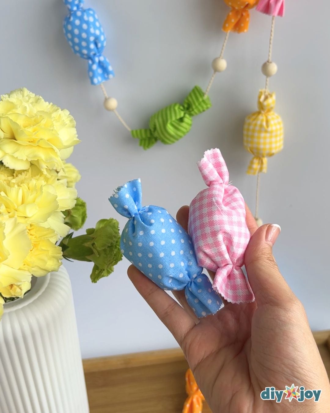 Hand holding a blue polka dot and a pink gingham fabric candy in front of a hanging no-sew garland and yellow carnations