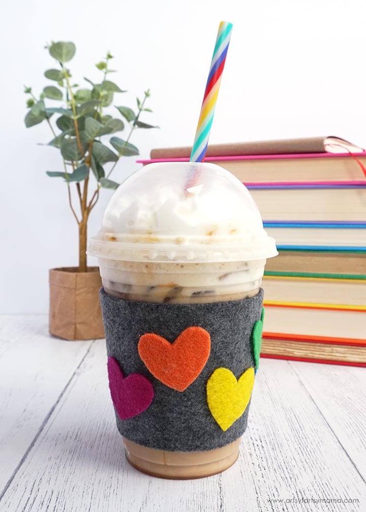 Gray felt cup cozy with pink, orange and yellow hearts wrapped around an iced coffee with whipped cream and a rainbow straw