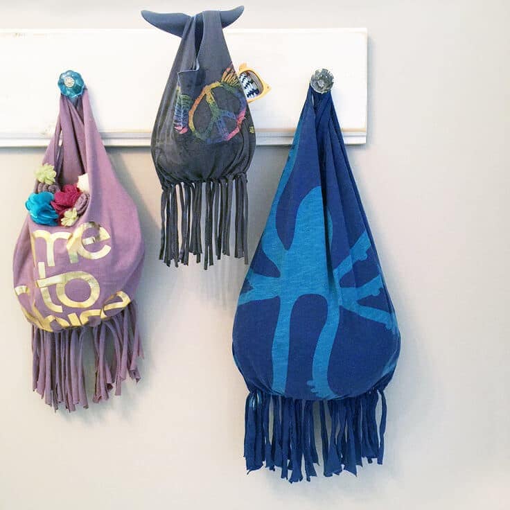 Three no-sew fringed T-shirt totes in purple, gray and blue hanging on knobs, one with fabric flowers and one with sunglasses