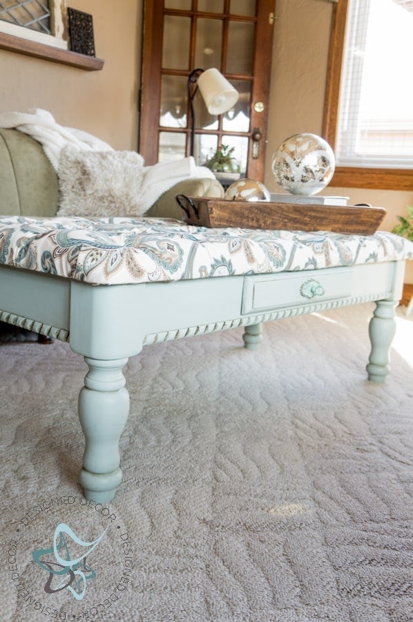 Coffee table bench with a diamond tufted floral fabric top and painted mint green legs on a carpet in a cozy living room
