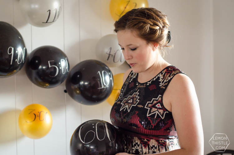 Woman holding a black balloon marked 6:00 in front of a wall of black, gold and white hourly countdown balloons