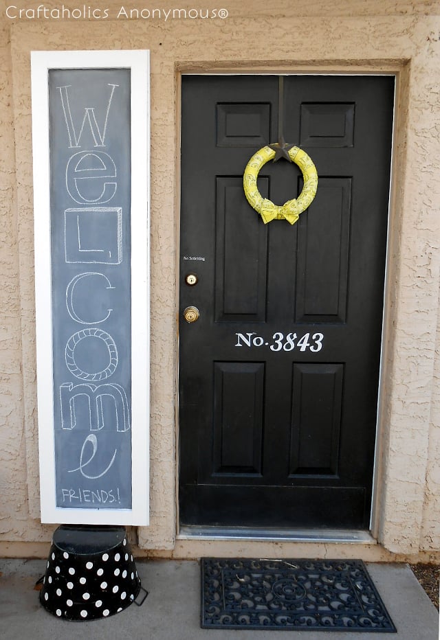 Tall narrow outdoor chalkboard with a white frame and Welcome Friends written in chalk, beside a black front door and bench