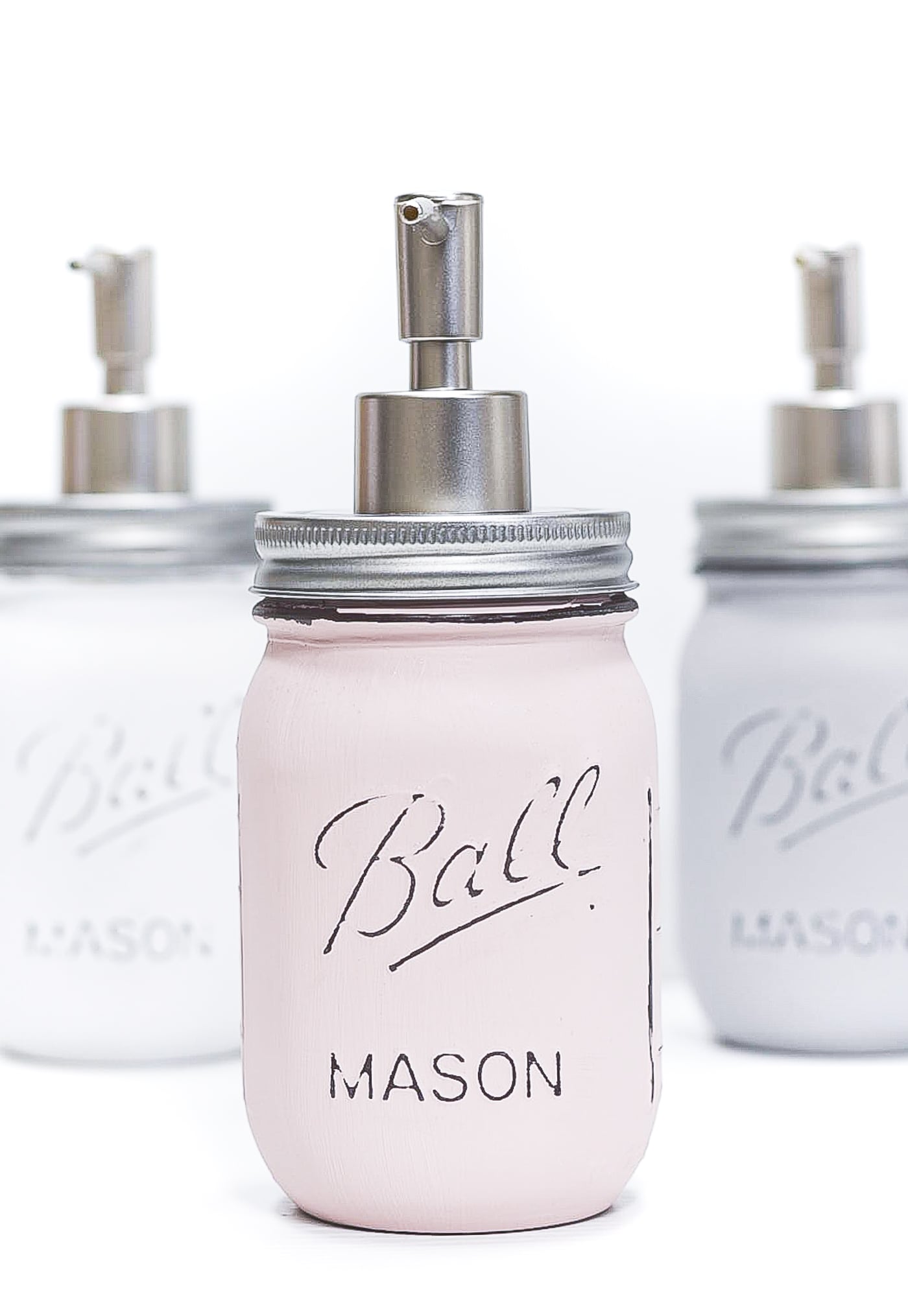 Three painted mason jar soap dispensers in blush pink, white and gray with distressed Ball lettering and silver pumps