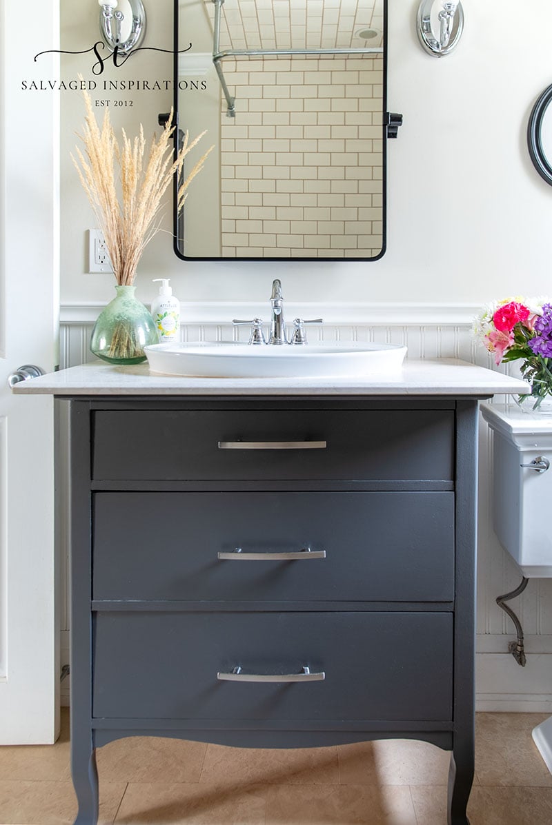 Charcoal gray painted bathroom vanity with three drawers and silver pulls under a white sink and black framed mirror