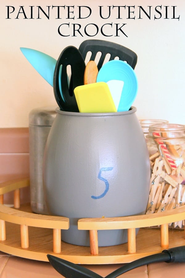 Painted farmhouse utensil crock in matte gray with a blue number 5, holding spatulas on a wood lazy Susan