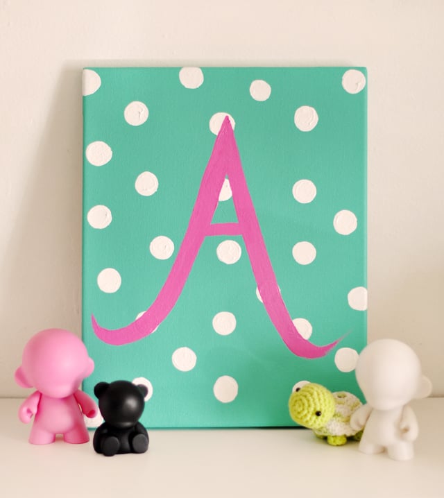 Teal canvas with white polka dots and a pink script letter A on a shelf with small vinyl toy figures and a crochet turtle