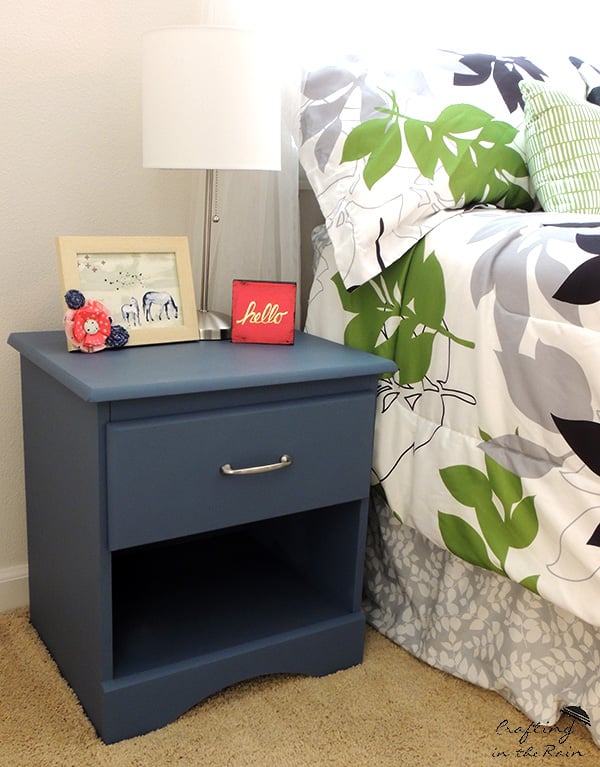 Navy painted laminate nightstand with a silver drawer pull beside a bed, topped with a white lamp and small framed photos