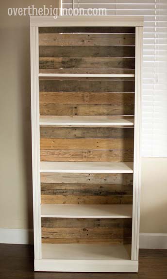 Tall white bookshelf with empty shelves and a back panel of mixed reclaimed pallet wood boards in warm and gray tones
