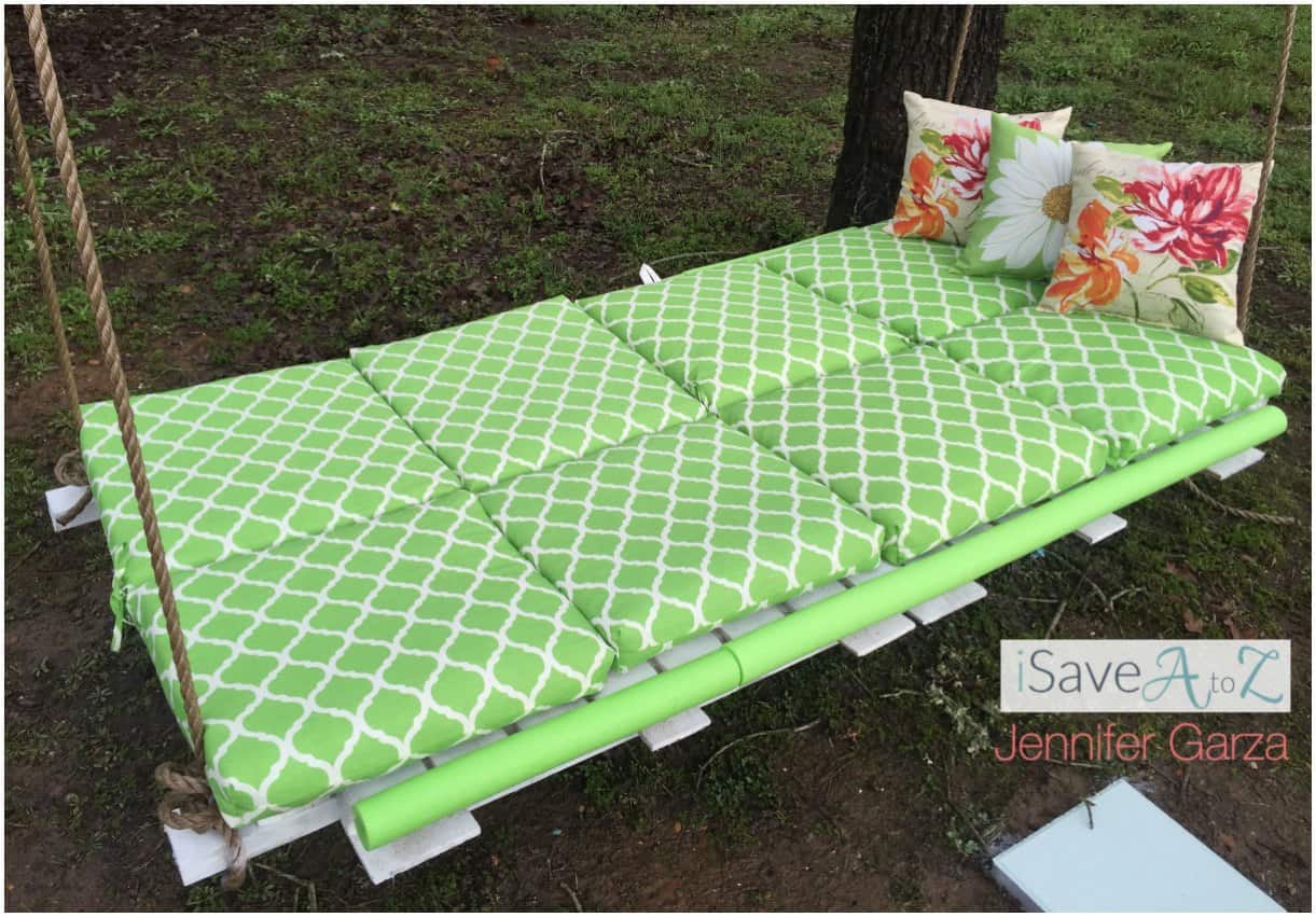 White pallet bed swing hung by rope from a tree, topped with green lattice print cushions and two floral outdoor pillows