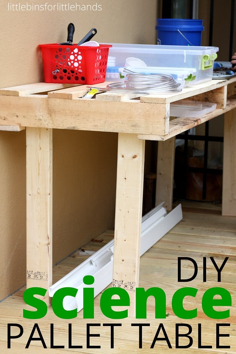 Pallet and 2x4 outdoor kids table on a deck holding a red basket, plastic bins and a blue bucket, with a lower slat shelf