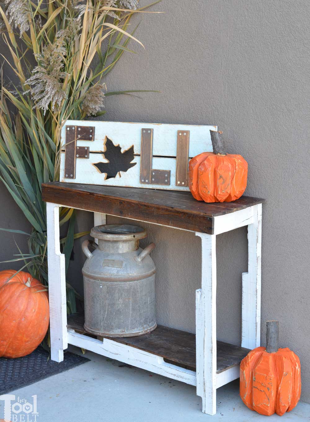 Pallet porch table with a white painted base and stained plank top, holding orange wood pumpkins beside an old milk can