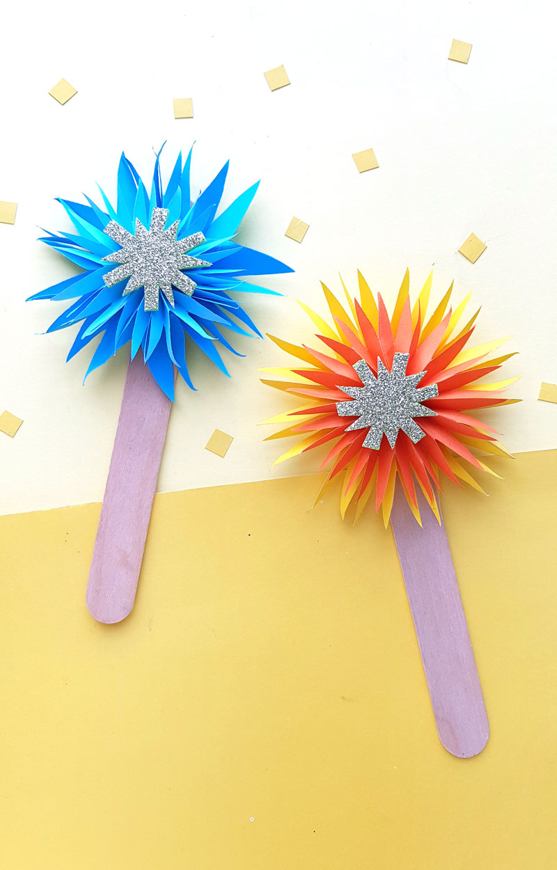 Blue and orange paper starburst fireworks with silver glitter star centers on purple popsicle sticks on yellow