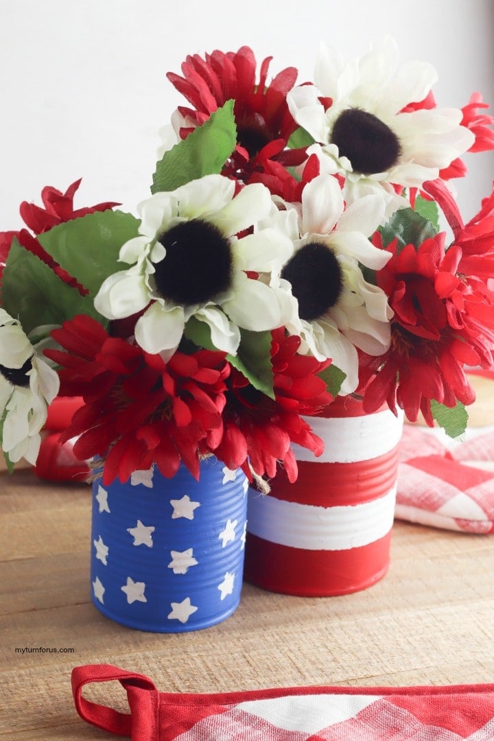 Two painted tin cans, blue with white stars and red and white stripes, holding red and white faux flowers